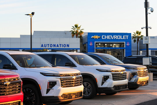 A Chevrolet dealership is seen at sunrise, with a lineup of new SUVs parked in front. The vehicles reflect golden light, and the AutoNation sign and Chevrolet showroom entrance are visible in the back