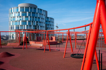 Open playground with red equipment and sculpted rubber flooring, modern buildings behind, ideal for active lifestyles and outdoor childhood design in Copenhagen, Denmark.