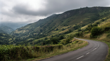 Naklejka premium A winding asphalt road snakes through rolling green hills under a cloudy sky, creating a scenic and peaceful journey through the natural landscape of mountains.