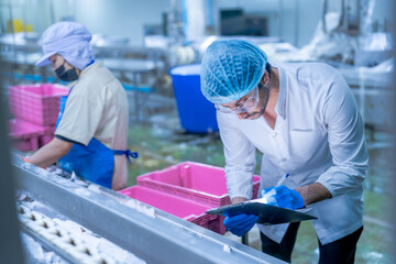 Workers in a food processing plant wearing protective gear, sorting fish on a conveyor belt into...