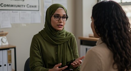 A woman in a green hijab listens intently as another woman shares her thoughts during a private conversation in a community center, fostering a sense of trust and support.