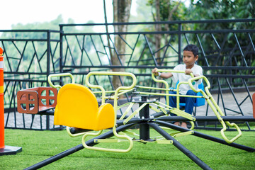 A young asian boy enjoys riding a colorful merry-go-round on a bright day at the outdoor playground.