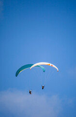 A pair of paragliding athletes are floating in the air showing their skills in controlling their paraglider against a backdrop of bright blue sky.