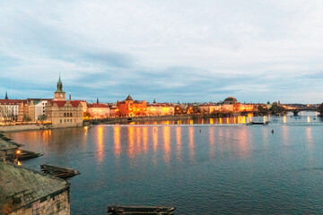 Naklejka premium A cityscape by the riverbank at dusk. The many lights of the city's buildings reflect in the river water, creating a warm and glowing effect.