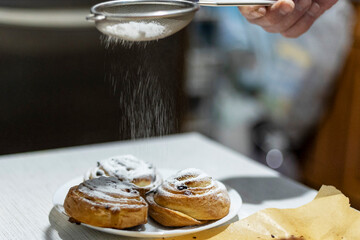 A hand dusts powdered sugar through a sieve over freshly baked cinnamon rolls in a kitchen setting....