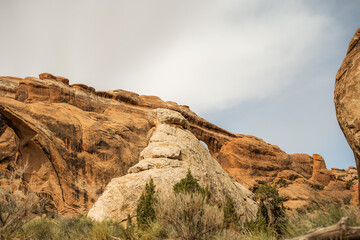 A prominent rock spire and distant sandstone arch rise from the desert terrain in Utah, surrounded by scrub vegetation and rugged red rock formations