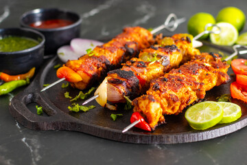 Grilled chicken skewers or chicken tikka kebab on a black plate with sliced tomatoes, onions, lime, green chutney, and ketchup, served on a dark marble background.
