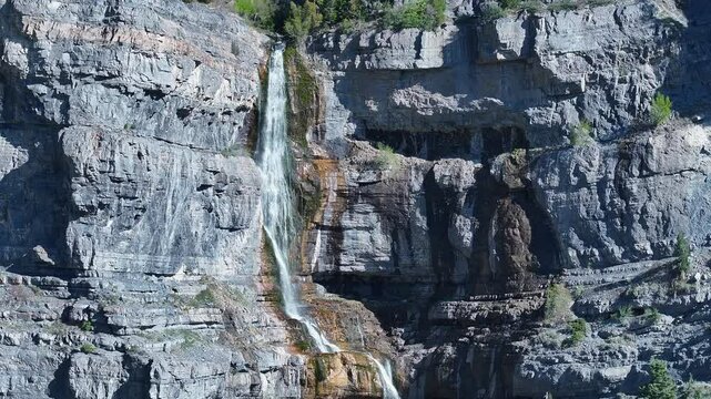 Aerial mountain Bridal Veil Falls Provo Utah 2 descend. 607 foot tall double cataract waterfall in Provo Canyon. Rugged rocky ledges and cliffs over pine forests. Deep canyon valley. Summer.