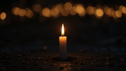 A close-up shot of a single, burning candle placed on dark ground, with a blurred background of out-of-focus warm golden lights.