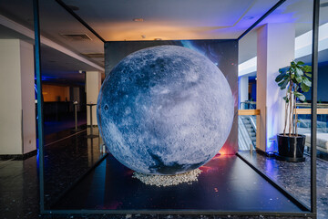 Indoor space installation with a large moon replica placed on a dark platform surrounded by stones, with space-themed decor in a modern building..