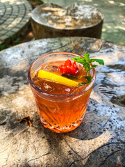 Vertical shot of a cocktail in a glass, garnished with a slice of lemon and a cherry, presented on a textured stone table, with natural sunlight.