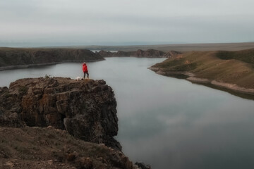 A person in a red jacket stands with a dog on a rocky cliff overlooking Kurtinskoye Reservoir near...