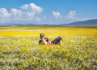 A female tourist lies with her Corgi dog in a field of yellow and white flowers against a backdrop of mountains and blue sky in the steppe of Kazakhstan