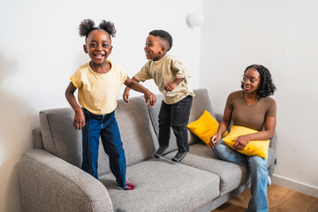 African two cheerful children are jumping on a grey sofa while their mother is smiling and watching them in their modern living room