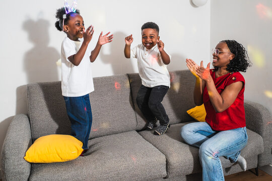 African cheerful mother watching her children jumping on the sofa, clapping hands and smiling, enjoying family time together