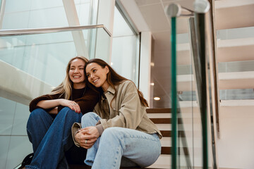 Fototapeta premium A blonde European woman in her early 20s, dressed in a black sweater, sits with a dark-haired Caucasian adult female friend and laughs in the hall.