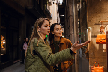 Walking down a narrow street while shopping, a blonde European woman in her early 30s shows off one bag and talks to her dark-haired female young friend in a brown jacket.