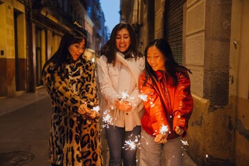 Fototapeta premium A dark-haired Caucasian woman in her early 20s, a European adult woman, and an Asian young woman wearing in warm jackets holding lit sparklers and smiling in a narrow street in the evening.