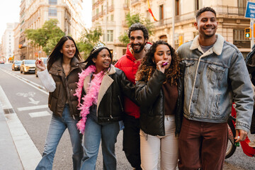 On a sunny street, a happy dark-haired Black birthday girl in her early 20s with a crown on her head walks and celebrates with a group of smiling multi-ethnic young friends.