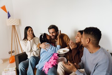 A dark-haired Black birthday girl in her early 20s kisses a cheerful dark-haired man in his early 30s with dark skin on the cheek while multi-ethnic adult friends look on in the living room.