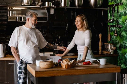 Standing in a modern kitchen, a smiling dark-haired European man in his early 40s wearing a white t-shirt talks and cooks with a blonde European woman at the table.