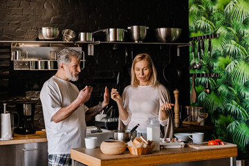 A European man in his 60s with short gray hair talking with a White wife in her late 50s while they are standing at a table with eggs, bread, and tomatoes in the kitchen with a black brick wall
