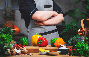 Chef confidently holds arms crossed over chest. Cooking food in the kitchen. with wooden table, kitchenware, vegetables, herbs and ingredients. Healthy eating, recipes, food blogging