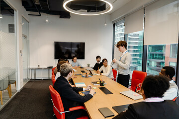 Side view of a dark-haired European man in his early 20s talking about a presentation in a meeting room while a group of multi-ethnic adult colleagues sit at a table in daylight.