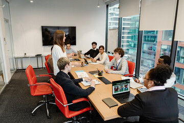 In a meeting room, a dark-haired European woman in her early 20s wearing a white shirt presents a report while standing near a table to multi-ethnic adult colleagues during the daytime.