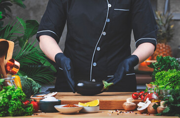 From farm to kitchen. Chef holds fresh eggplant in hands. Wooden table, kitchenware, vegetables, herbs and ingredients. Healthy vegan eating, recipes, blogging