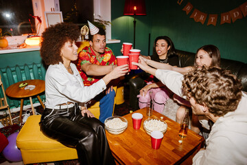 A multiethnic group of friends in their late 20s consisting of three women and two men sitting around a table and toasting with red cups of drinks during a birthday party in a room with green walls