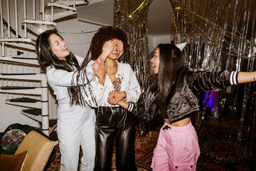 Side view of a joyful dark-haired Asian woman in her early 20s closing the eyes of her curly-haired Black adult friend while the dark-haired adult woman holds her hand at a party in the living room.