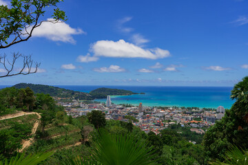Naklejka premium Ultra-sharp midday vista from a jungle ridge above Patong Bay, Thailand: emerald palms and terraced hills frame turquoise water, a cobalt sky with cotton clouds, and a cluster of modern towers and low