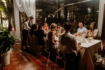 A back view of a dark-haired adult man talking to a smiling European woman in her early 20s in a gold dress while other multi-ethnic guests celebrate a wedding in the restaurant.