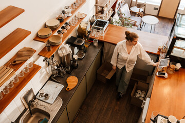 A serious, dark-haired European woman owner in her early 30s in a white shirt holding a box in one...