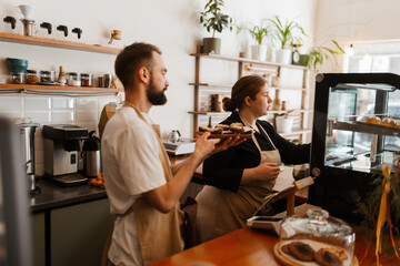 Side view of a dark-haired adult male barista in a white t-shirt holding desserts while an attentive female European owner in her early 30s puts them in the fridge at the coffee shop.