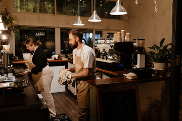 Side view of a dark-haired man European barista in his early 30s, wiping a plate and looking at a...