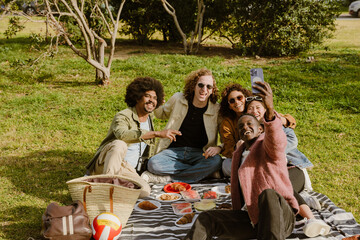 A Black man in his 20s taking a selfie with a multiethnic, group of young adult friends while they are sitting on a blanket scattered on the grass in a park during a picnic with plates of various food