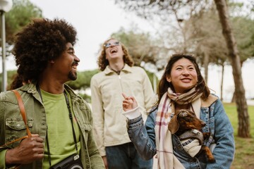 Fototapeta premium A happy Asian woman in her early 20s holding a dog on her arm, with her other hand poking at multi-ethnic young friends laughing while walking in a park.
