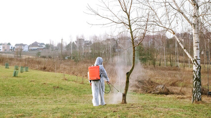 Gardener in protective clothing with a knapsack sprayer is spraying bush and tree with fungicide in a spring garden. Plant protection from diseases, insects and pests control. High quality photo.