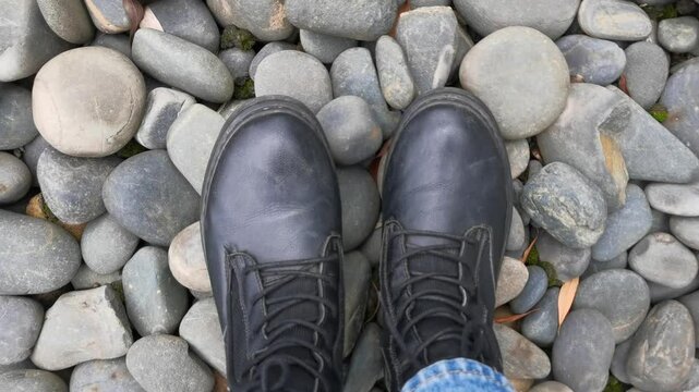 POV top view leg in black Short boots under the ground covering pepples or stones.Shotting a pair of black boots from up to down after walking