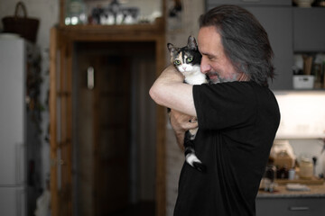 An adult man with gray hair against the background of a home interior with a cat in his arms. He hugs her and smiles.