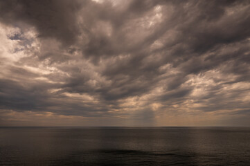 Fototapeta premium Sun between dark storm clouds over ocean. Dramatic seascape. Clouds and rain flurry over sea.