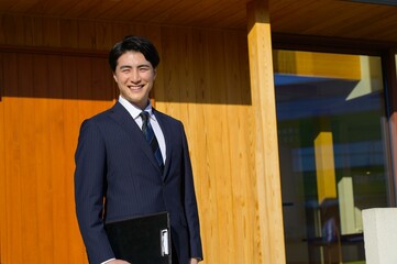 スーツ姿の男性が玄関先で明るく立っているプロフェッショナルな印象の一枚 © Yamatatsu