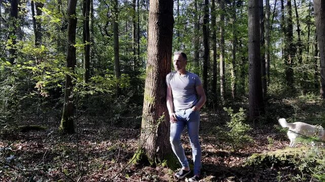 Attractive fit sporty slim man intight jeans and muscle top in thoughtful pose leaning against tree in summer woodland with dog in background