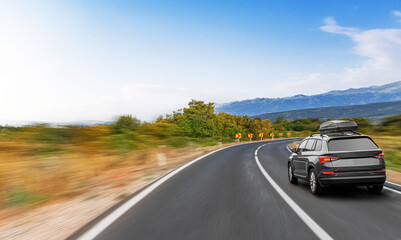 A modern SUV with a rooftop cargo box drives along a winding country road surrounded by trees and mountains under a clear blue sky.