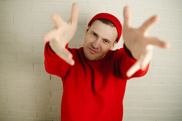 A man in a red hoodie and hat dances against a white brick wall