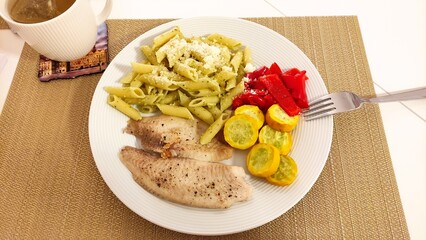 Grilled fish fillets are served alongside pesto pasta, roasted yellow squash, and red bell peppers,...