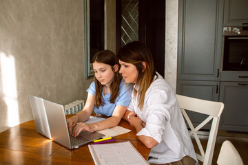 Mother and daughter using laptop sitting together in home kitchen. Mom helping kid with school...