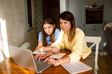 Mother and daughter using laptop sitting together in home kitchen. Mom helping kid with school...